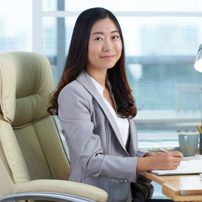 Cheerful Asian woman sitting at desk in office, writing and posing for camera